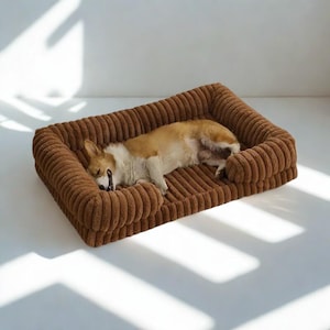 May include: A brown corduroy dog bed with a sleeping corgi. The bed has a rectangular shape with raised sides and a soft, textured surface. The dog is light brown and white, curled up on the bed.
