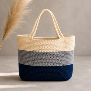 May include: A tote bag featuring a color-blocked design with three horizontal sections. The top section is cream, the middle section is light gray, and the bottom section is dark navy blue. The bag has two integrated handles and a woven texture.