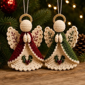 May include: Two crocheted angel ornaments with gold halos, one in deep red and one in forest green. Each figure features cream-coloured wings with embroidered details, joined hands, and a holly leaf and berry embellishment on the front hem.
