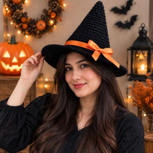 May include: A black crocheted witch hat featuring a pointed crown and a wide brim. The hat is accented with a vibrant orange satin ribbon tied into a bow around the base of the crown. The texture of the hat shows visible yarn stitches.