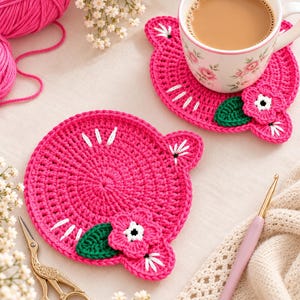 May include: Two round, hot pink crocheted coasters featuring white stitched details, a small green leaf, and a pink flower with a white center. One coaster holds a white ceramic mug with a floral pattern, while the other sits near gold scissors.