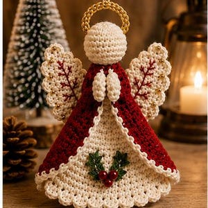 May include: A crocheted angel ornament featuring a cream-colored head and base, a deep red cloak, and a gold halo. The wings are cream with red stitching and small gold beads. A holly sprig with three red berries is centered on the front.