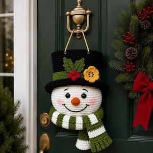 Puede incluir: Un adorno de puerta de ganchillo con forma de muñeco de nieve que lleva un sombrero de copa negro con una banda verde, hojas verdes, bayas rojas y una flor amarilla. El muñeco tiene cara blanca, ojos negros, nariz naranja y una bufanda a rayas.