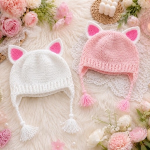 May include: Two crochet beanies with cat ears, one in solid white and one in light pink. Both hats feature bright pink inner ear details, ear flaps, and braided ties ending in matching yarn tassels. The construction shows a dense stitch pattern.