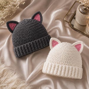 May include: Two crocheted beanies, one dark grey and one off-white, each featuring a ribbed brim and two pointed cat ears with pink inner detailing. The hats are constructed with a dense, textured stitch pattern and have a folded cuff design.