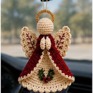 May include: A crocheted angel ornament hanging from a string, featuring a cream-colored head and halo, a deep red dress with cream trim, and wings with red stitching. The front of the dress is decorated with a small green holly and red berry motif.