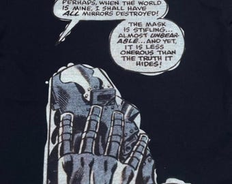 May include: A black t-shirt featuring a graphic print of a metallic mask held by a hand. The print includes comic book speech bubbles with the text: PERHAPS, WHEN THE WORLD IS MINE, I SHALL HAVE ALL MIRRORS DESTROYED! THE MASK IS STIFLING... AND BAH! ENOUGH OF THIS!