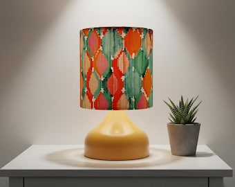 May include: A yellow table lamp with a patterned lampshade featuring a design of colorful, overlapping shapes in shades of orange, green, pink, and beige. The lamp sits on a white nightstand next to a small potted plant.