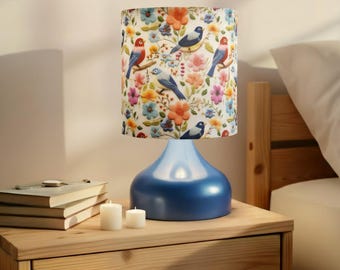May include: A blue ceramic table lamp with a floral and bird-patterned lampshade. The lampshade features colorful flowers and birds in shades of blue, red, and yellow. The lamp is on a wooden nightstand with books and candles.