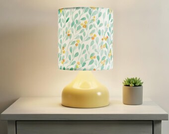 May include: A yellow table lamp with a cylindrical lampshade featuring a watercolor pattern of green leaves and yellow berries. The lamp sits on a white nightstand next to a small potted succulent.