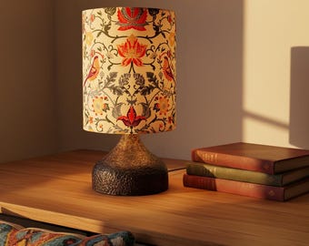 May include: A decorative table lamp with a patterned lampshade featuring red, orange, and black floral and bird designs. The lamp has a textured, dark brown base and sits on a wooden surface, next to a stack of books.