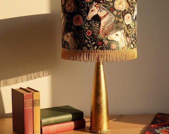 May include: A decorative table lamp with a gold base and a lampshade featuring a horse and floral design. The lampshade has a black background with images of white horses, pink flowers, and green foliage. The lamp is next to a stack of books.