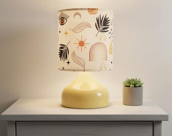 May include: A cream-colored table lamp with a patterned lampshade featuring suns, rainbows, leaves, and eyes. The base is a soft yellow color. A small potted succulent sits beside the lamp on a gray nightstand.