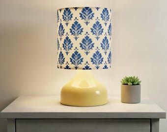May include: A yellow table lamp with a blue and white patterned lampshade sits on a white nightstand. A small succulent in a gray pot is next to the lamp. The lamp has a rounded base.