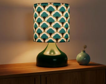 May include: A green glass table lamp with a cylindrical lampshade. The lampshade features a teal, gold, and black fan pattern. A small teal vase with a single stem sits to the right of the lamp on a wooden surface.