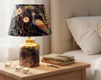 May include: A decorative table lamp with a dark shade featuring a peacock design. The lamp has a gold and brown base and sits on a wooden nightstand. Small candles and books are also on the nightstand.