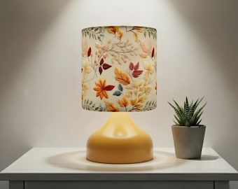 May include: A yellow table lamp with a floral lampshade featuring autumn leaves in shades of orange, red, and yellow. A small potted succulent sits beside the lamp on a white surface. The lamp base is a solid, rounded shape.