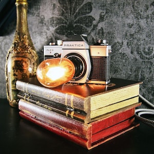 Könnte beinhalten: Eine Vintage Praktica PLC3 Kamera liegt auf einem Stapel antiker Bücher mit vergoldeten Seiten. Eine leuchtende Glühbirne und eine dekorative Flasche sind ebenfalls vorhanden. Die Szene hat eine warme, einladende Ästhetik.