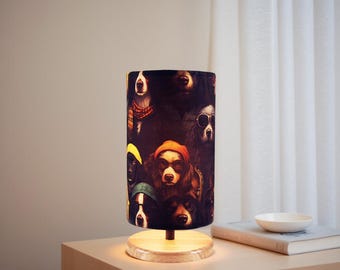 May include: A decorative table lamp with a cylindrical shade featuring a collage of dogs wearing hats and sunglasses. The lamp has a wooden base and is illuminated, casting a warm glow. The lamp is on a wooden nightstand.