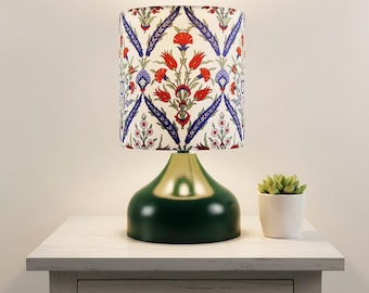 May include: A decorative table lamp with a green base and a patterned lampshade. The lampshade features a white background with red, blue, and green floral designs. The lamp sits on a white nightstand next to a small potted succulent.