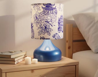 May include: A blue table lamp with a white lampshade featuring a blue floral and pagoda design. The lamp sits on a wooden nightstand with books and candles. The lamp base has a glossy finish.