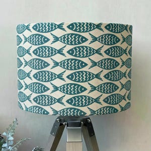 Puede incluir: Pantalla de lámpara cilíndrica con un patrón repetido de peces turquesa sobre un fondo crema. El diseño de los peces tiene un patrón en zigzag en el centro. La pantalla está montada sobre una base de lámpara trípode de madera.