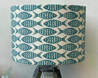May include: A cylindrical lampshade featuring a repeating pattern of teal fish on a cream background. The fish design has a zig-zag pattern in the center of the fish. The lampshade is mounted on a wooden tripod lamp base.