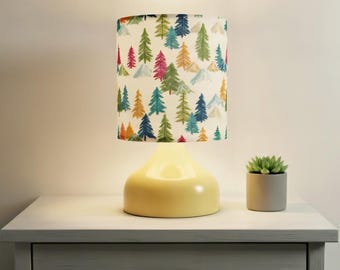 May include: A yellow table lamp with a cylindrical lampshade featuring a colorful forest pattern. The lamp sits on a white nightstand next to a small potted succulent. The lamp's base has a rounded shape.