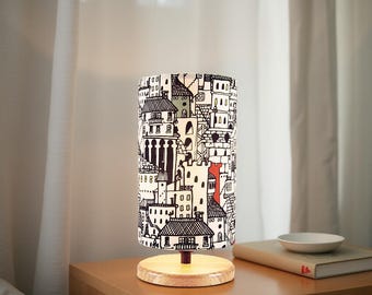 May include: A cylindrical table lamp with a city-themed lampshade. The lampshade features a black and white illustration of buildings with red accents. The lamp sits on a wooden base and is placed on a wooden nightstand.