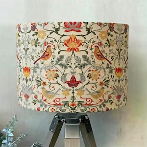 May include: A cylindrical lampshade featuring a cream background with a repeating pattern of birds, red and yellow flowers, and green foliage. The design includes stylised botanical elements and avian figures arranged in a symmetrical layout.