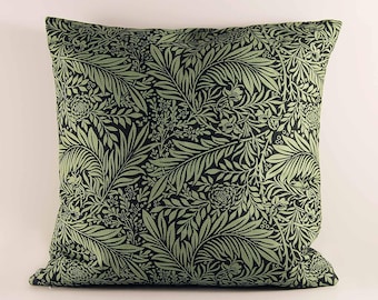 May include: A square throw pillow featuring a dense, repeating botanical pattern of sage green leaves, ferns, and small flowers set against a dark black background. The fabric has a visible woven texture, and the pillow has a square shape.