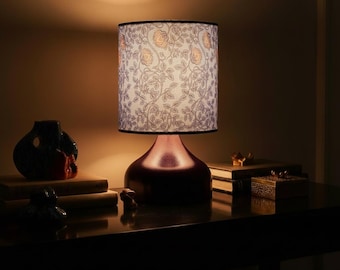 May include: A table lamp with a floral patterned lampshade. The base is a dark reddish-brown color. The lampshade has a white background with a floral design in shades of gray and orange. The lamp is on a dark wooden table.