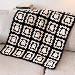 Peut inclure: Une couverture en crochet faite à la main présentant un motif répétitif de carrés. Chaque carré est composé d'une bordure noire entourant un centre crème représentant une silhouette de fantôme stylisée avec deux yeux sombres.