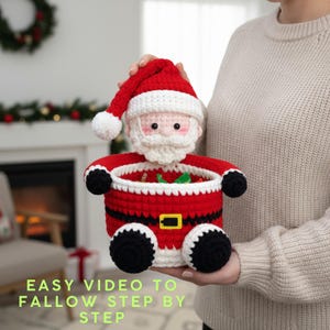 Peut inclure: Un panier fait main en crochet représentant le Père Noël, rouge, blanc, noir et beige. Le panier a un bonnet, un visage et une barbe de Père Noël. Le texte "EASY VIDEO TO FOLLOW STEP BY STEP" est en bas de l'image.