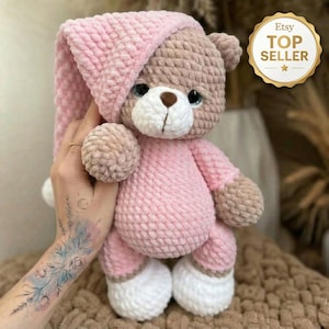 Peut inclure: Un ours en peluche crocheté, vêtu d'un bonnet de nuit et d'un pyjama roses. L'ours a un visage, des bras et des jambes marron clair, avec des accents blancs sur les pieds. Un badge Etsy Top Seller est visible.