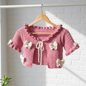 May include: A pink, short-sleeved crochet cardigan with white bow accents. The cardigan has a ruffled collar and sleeve edges, and a tie closure. The garment is displayed on a wooden hanger against a white brick wall.