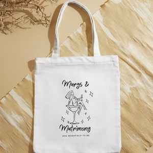 May include: White tote bag with black text and illustration. The text reads "Margs & Matrimony" with a drawing of a person in a martini glass. Below the text is "MRS WAKEFIELD TO BE". The bag has two white handles.