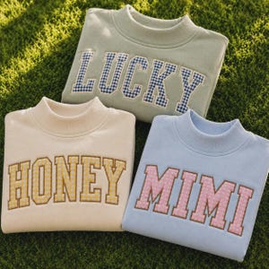 May include: Three crewneck sweatshirts in sage green, cream, and light blue. The sage green sweatshirt features the word LUCKY in blue gingham letters. The cream sweatshirt says HONEY in yellow gingham, and the light blue one says MIMI in pink.