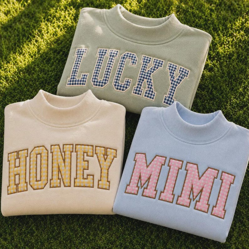 May include: Three crewneck sweatshirts in sage green, cream, and light blue. The sage green sweatshirt features the word LUCKY in blue gingham letters. The cream sweatshirt says HONEY in yellow gingham, and the light blue one says MIMI in pink.
