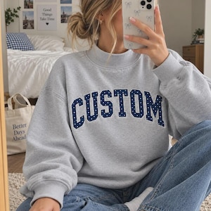 May include: A light gray crewneck sweatshirt featuring the word CUSTOM in dark blue block letters with white polka dots. The sweatshirt has long sleeves, a ribbed collar, and a relaxed fit, worn with blue denim jeans.