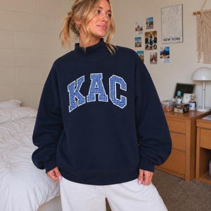 May include: A navy blue mock neck sweatshirt featuring the letters KAC in a white and blue gingham check pattern across the chest. The garment has long sleeves with ribbed cuffs and a ribbed hem, providing a relaxed fit.