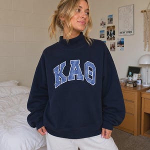 May include: A navy blue long-sleeve sweatshirt with a mock neck collar. The front features the Greek letters KAO in a white and blue gingham check pattern with a white outline, arranged in an arched design across the chest area.