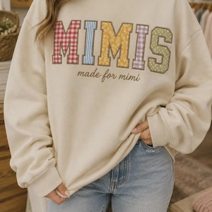 May include: A cream-coloured mock neck sweatshirt featuring the word MIMIS in large, multi-coloured patterned fabric letters, with the text made for mimi embroidered in brown cursive script underneath the main lettering on the front.