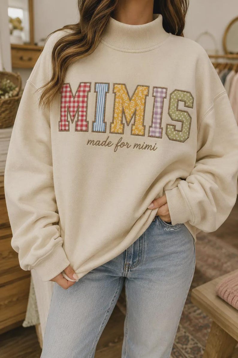 May include: A cream-coloured mock neck sweatshirt featuring the word MIMIS in large, multi-coloured patterned fabric letters, with the text made for mimi embroidered in brown cursive script underneath the main lettering on the front.