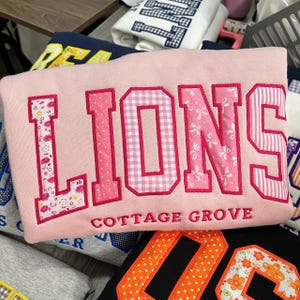 May include: A light pink sweatshirt featuring the word LIONS in large, block-style applique letters. Each letter has a unique pink pattern, including floral, polka dot, gingham, bow, and striped prints. The text COTTAGE GROVE is embroidered below.