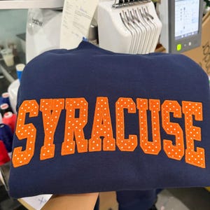 May include: A navy blue sweatshirt featuring the word SYRACUSE in large, block-style letters. The letters are orange with a white polka dot pattern and are appliqued onto the front of the garment in a slightly arched arrangement.