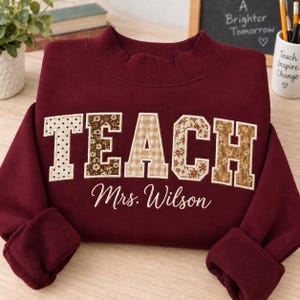 May include: A maroon long-sleeve sweatshirt featuring the word TEACH in large block letters with varied patterns including polka dots, floral prints, and gingham. Below the word, the name Mrs. Wilson is embroidered in a white cursive script.