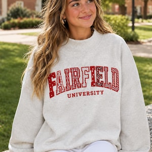 May include: A light grey crewneck sweatshirt featuring the word FAIRFIELD in large, red, patterned block letters with various textures, and the word UNIVERSITY in smaller red letters underneath, all centred on the chest of the garment.
