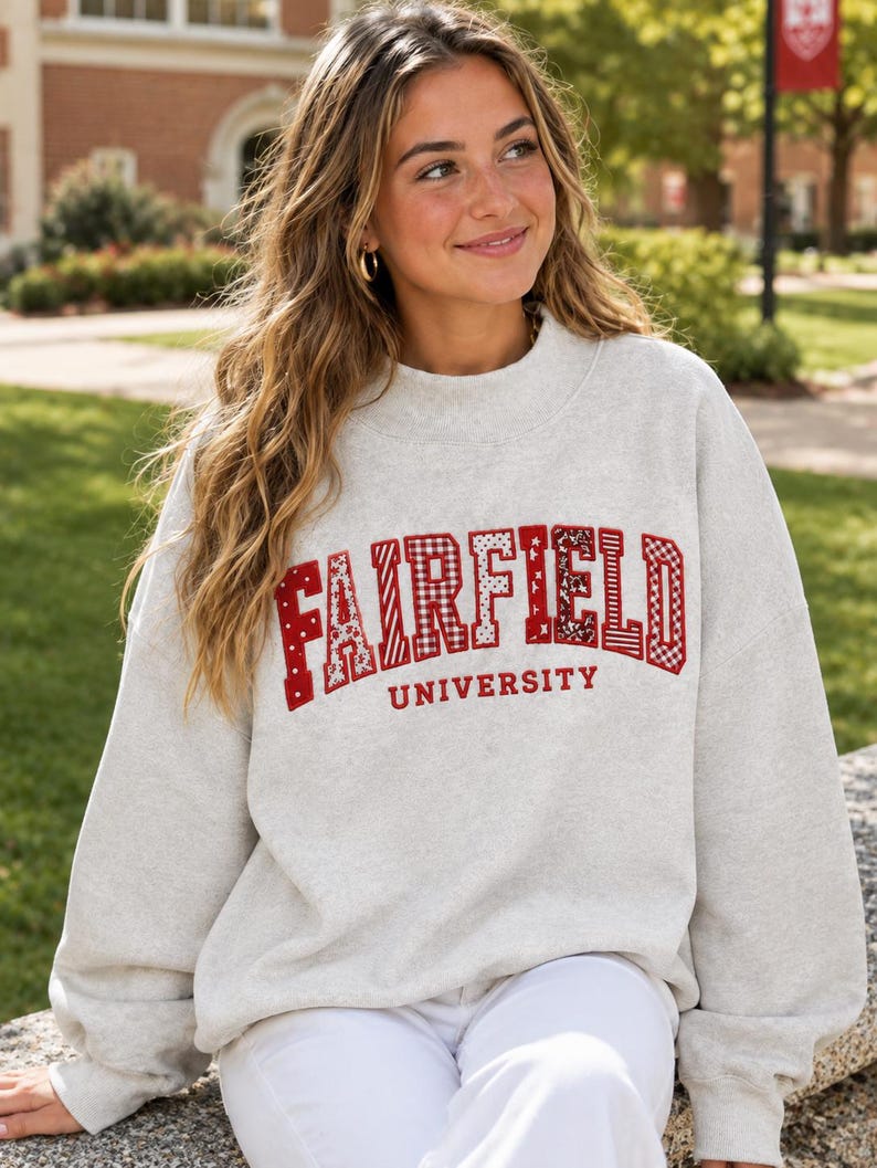 May include: A light grey crewneck sweatshirt featuring the word FAIRFIELD in large, red, patterned block letters with various textures, and the word UNIVERSITY in smaller red letters underneath, all centred on the chest of the garment.