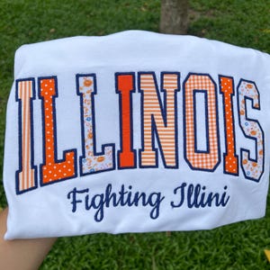 May include: A white t-shirt featuring the word ILLINOIS in large, block-style letters with varied orange and white patterns, including stripes, polka dots, and floral prints, outlined in navy blue. Below, the text Fighting Illini is embroidered.